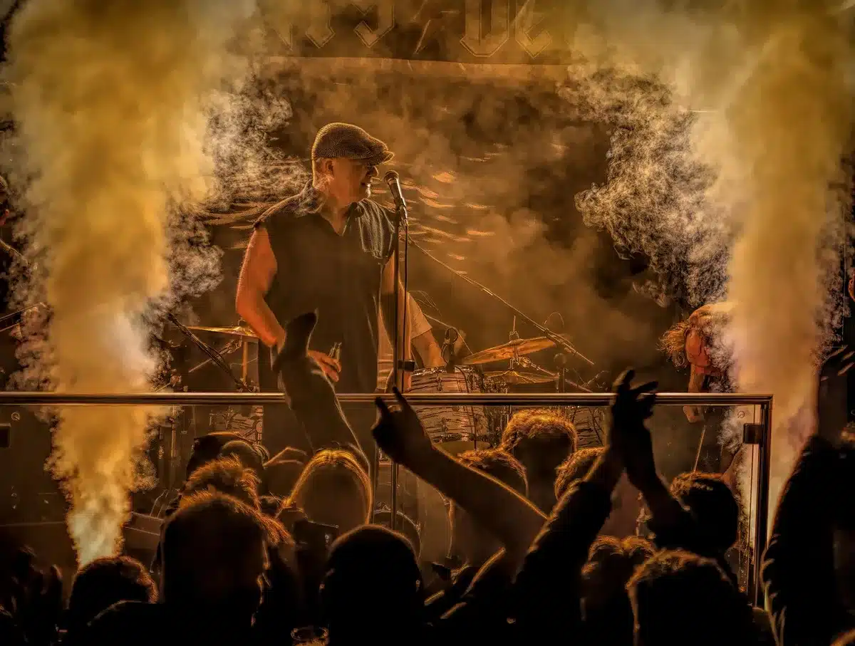 Concert stage with dramatic lighting, anonymous silhouettes of band members mid-performance against dark backdrop, crowd energy visible in distance, metalcore aesthetics