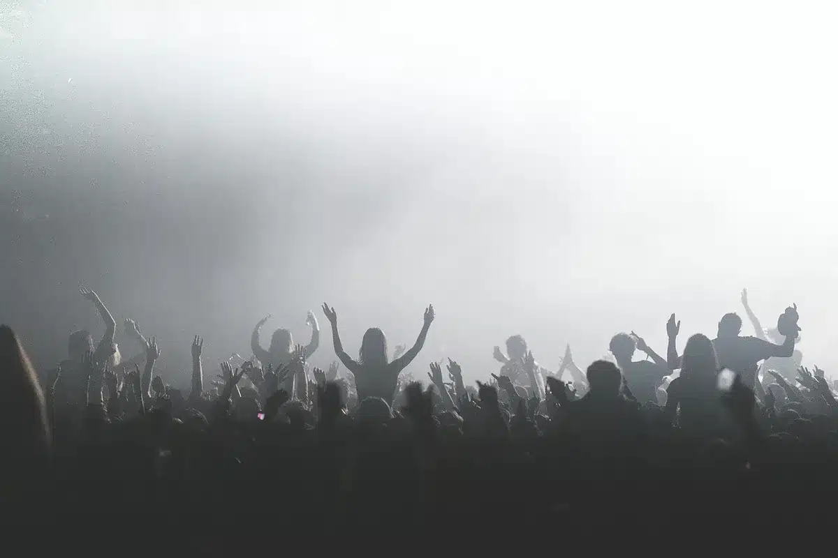A packed festival stage under bright floodlights, silhouettes of thousands of fans with raised arms and dancing bodies, energy and motion filling the massive outdoor venue, confetti or light effects in the air
