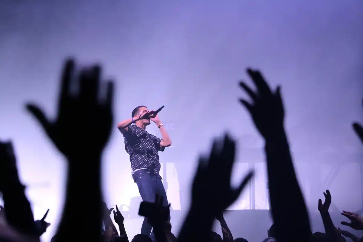 A spotlight illuminating a microphone on a darkened stage, with blurred silhouettes of performers in the background, electronic stage production elements visible, the energy of a live performance captured