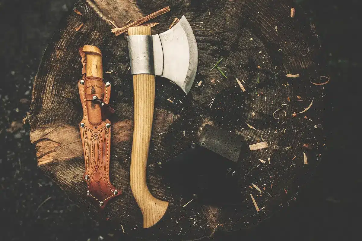 Two anonymous hands reaching toward a vintage axe on a wooden table, soft window light and period fabrics in the background