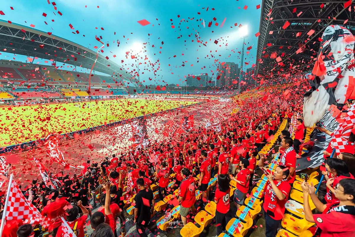Anonymous fans erupting in a stadium terrace, arms raised and scarves aloft, faces blurred in motion under floodlights, collective celebration and relief visible in silhouettes.