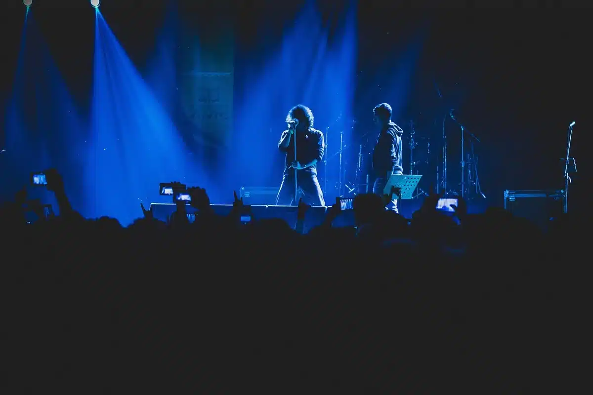 A packed festival stage at night with spotlights illuminating a rock band performing, thousands of silhouetted fans with raised arms and phone lights, festival tents and structures visible in the background, electric energy and movement frozen mid-performance