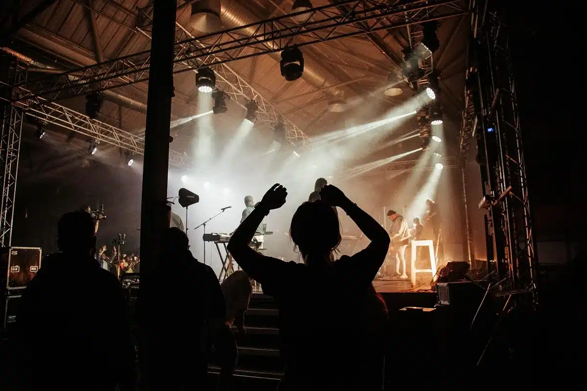 A close-up of a microphone stand under a single spotlight on an empty stage, stage lights blurred in the background, dust particles visible in the beam, coiled cables trailing into shadow