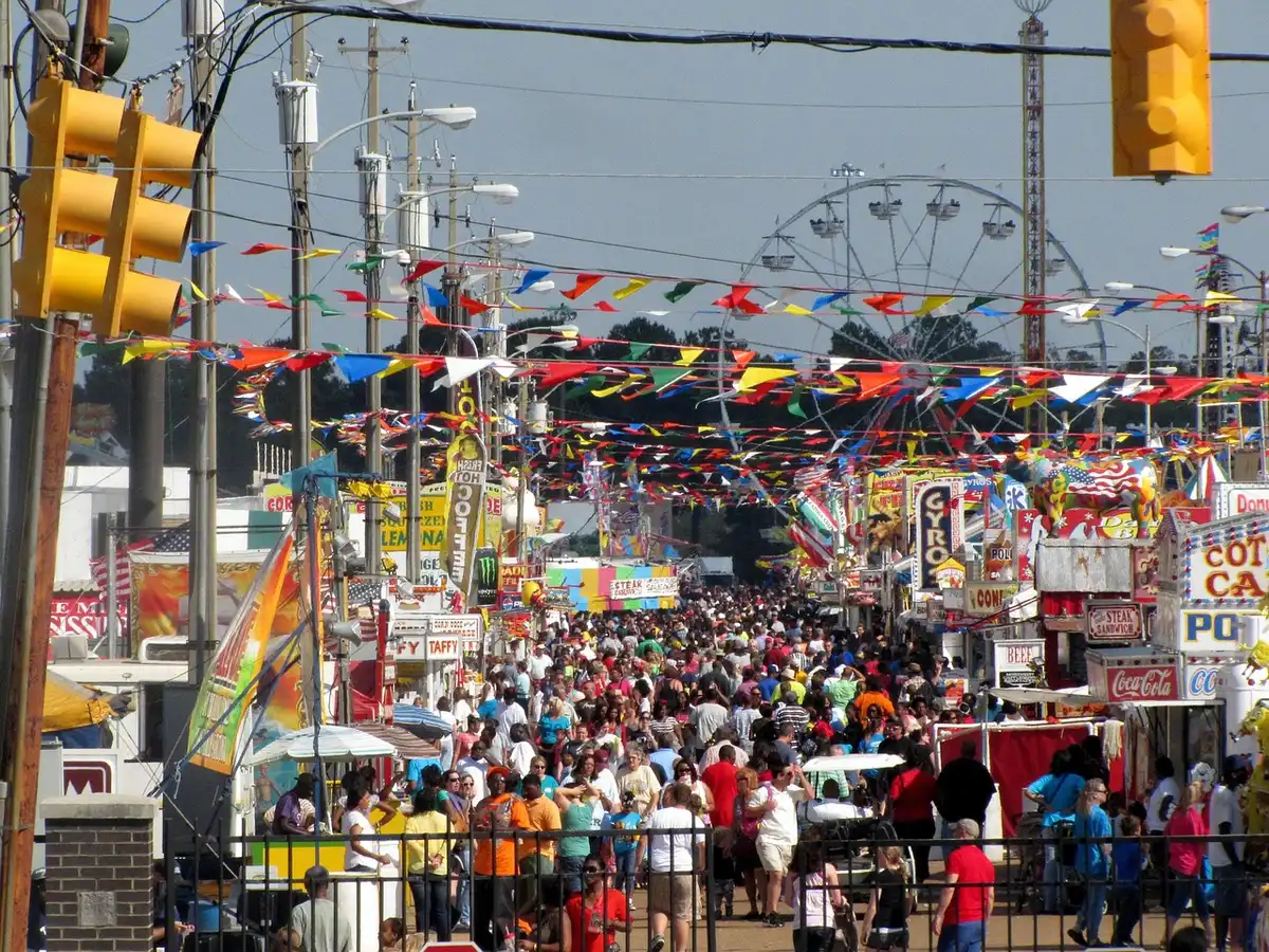 Crowded street carnival scene with metal barriers, dense mass of people pressed together, sense of confinement and urgency visible