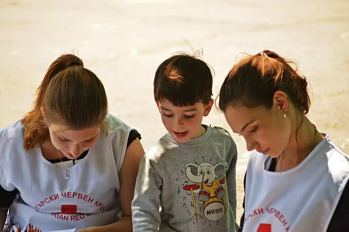 Four children standing together in a shelter interior, looking out a window toward a distant city skyline, expressions showing longing and displacement, soft institutional lighting.