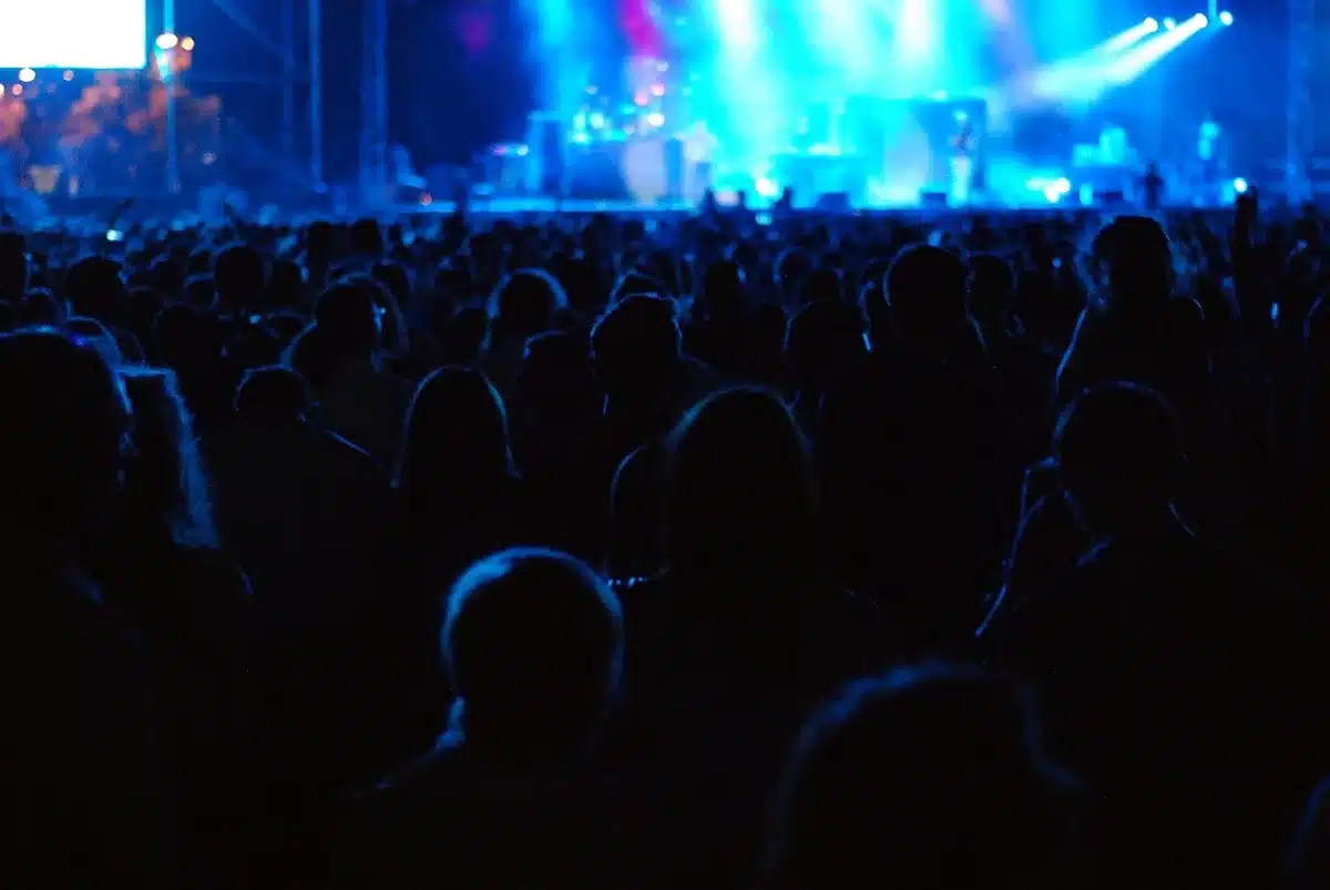 A packed festival arena at dusk with multiple stage structures illuminated, crowds of anonymous attendees under colorful stage lighting, diverse music fans celebrating together