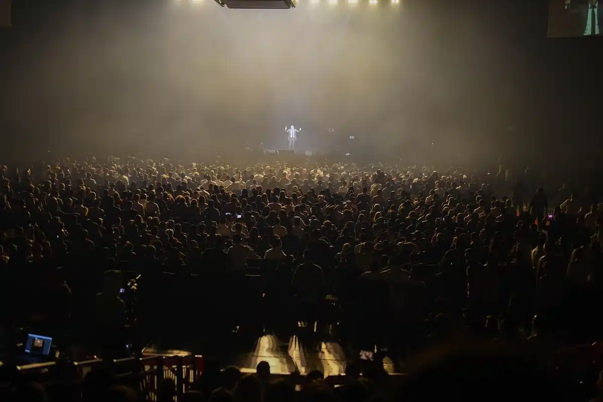 Massive outdoor stage with LED screens under evening lights, crowd silhouettes in distance, multiple performance areas visible across festival grounds