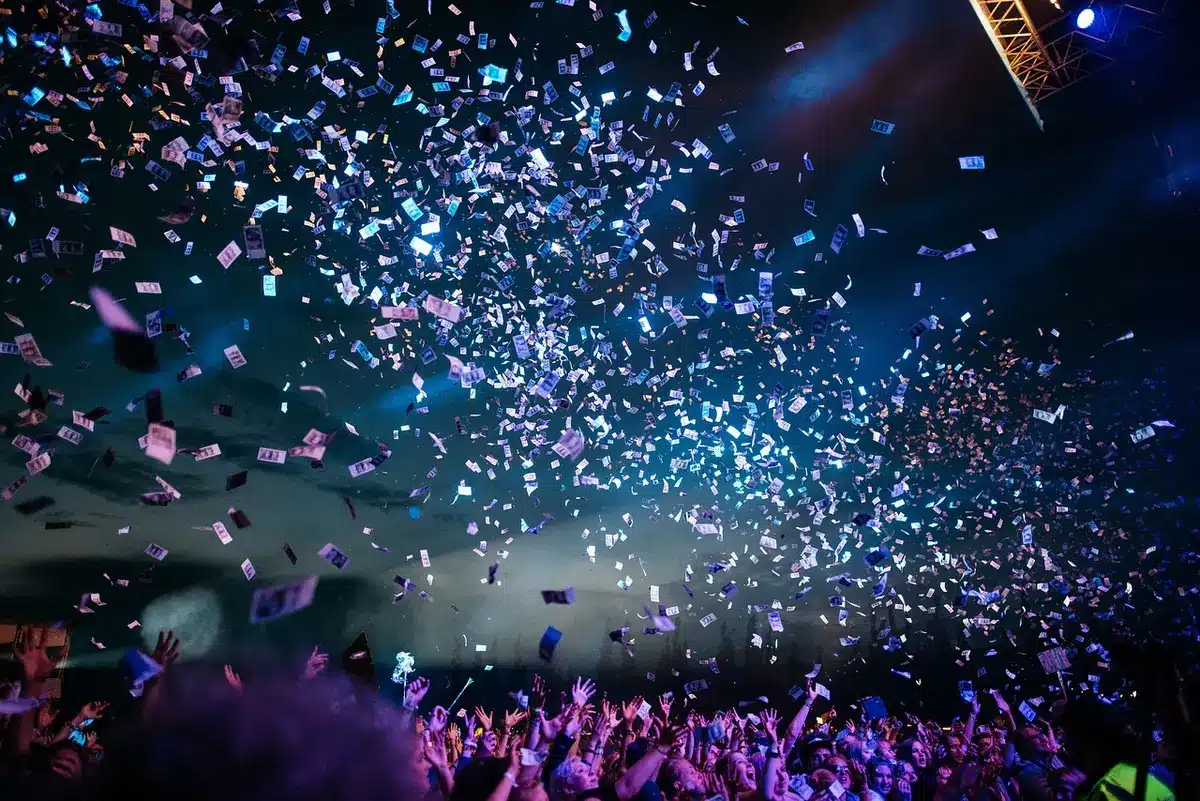 A concert stage with a glowing DJ booth and turntables, abstract neon lights reflecting off equipment, anonymous silhouettes of dancing hands in the foreground, the glow of smartphone screens in the crowd