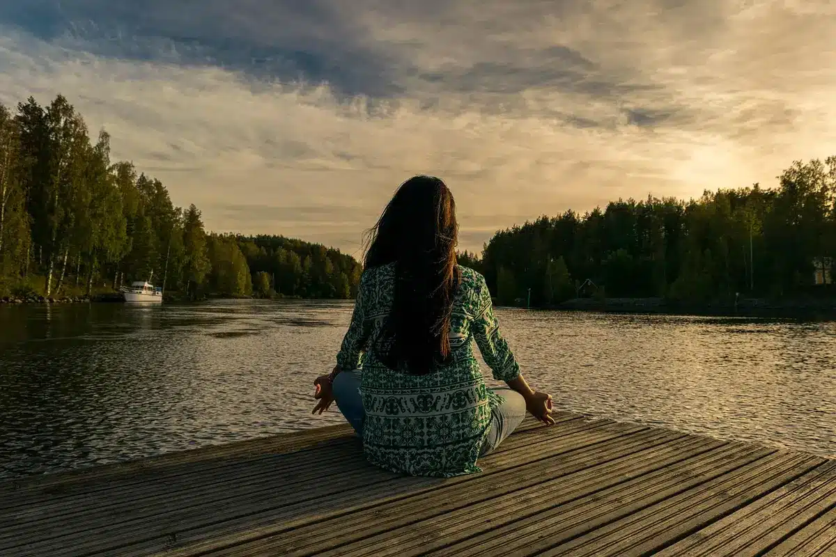A woman in her early 60s, sitting alone in a bright, sunlit room, hands clasped thoughtfully in her lap, a gentle expression of peace and contemplation on her face, soft natural light streaming through windows behind her, suggesting quiet reflection and personal growth.
