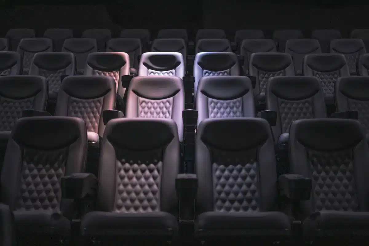 Empty cinema auditorium with a single lit screen and a few blurred anonymous silhouettes in seats, evoking the theatrical experience