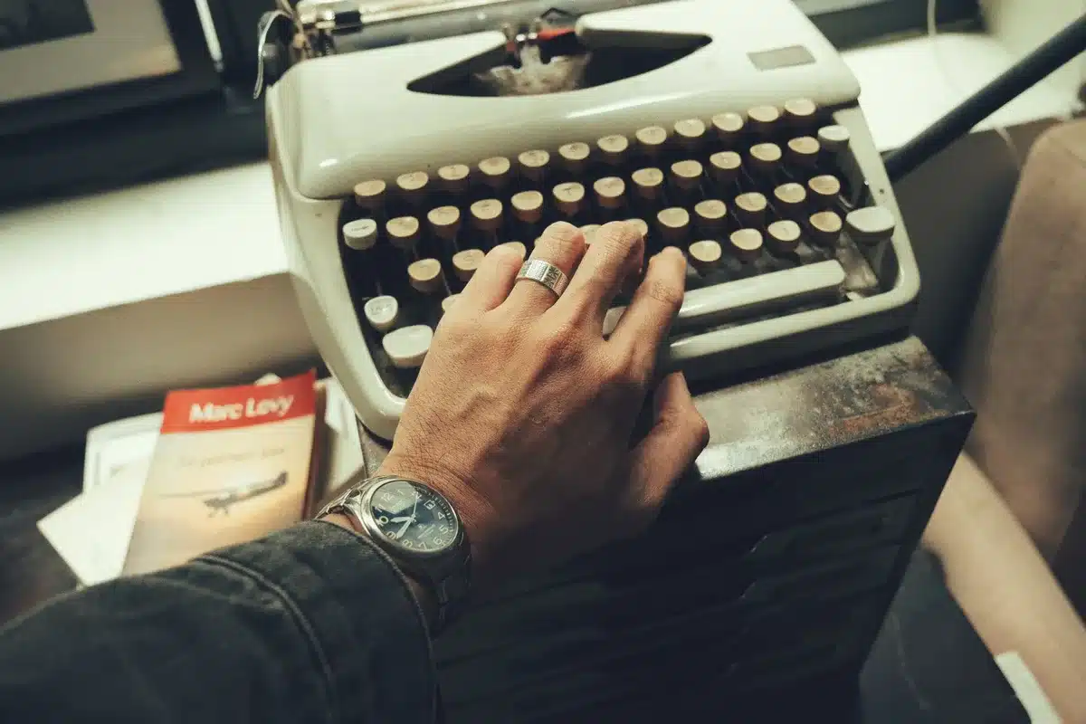 Empty wooden table with an open blank notebook, a pair of anonymous reading glasses resting beside it, and a single unbranded paperback stacked nearby — a quiet writer's desk suggesting imminent publication