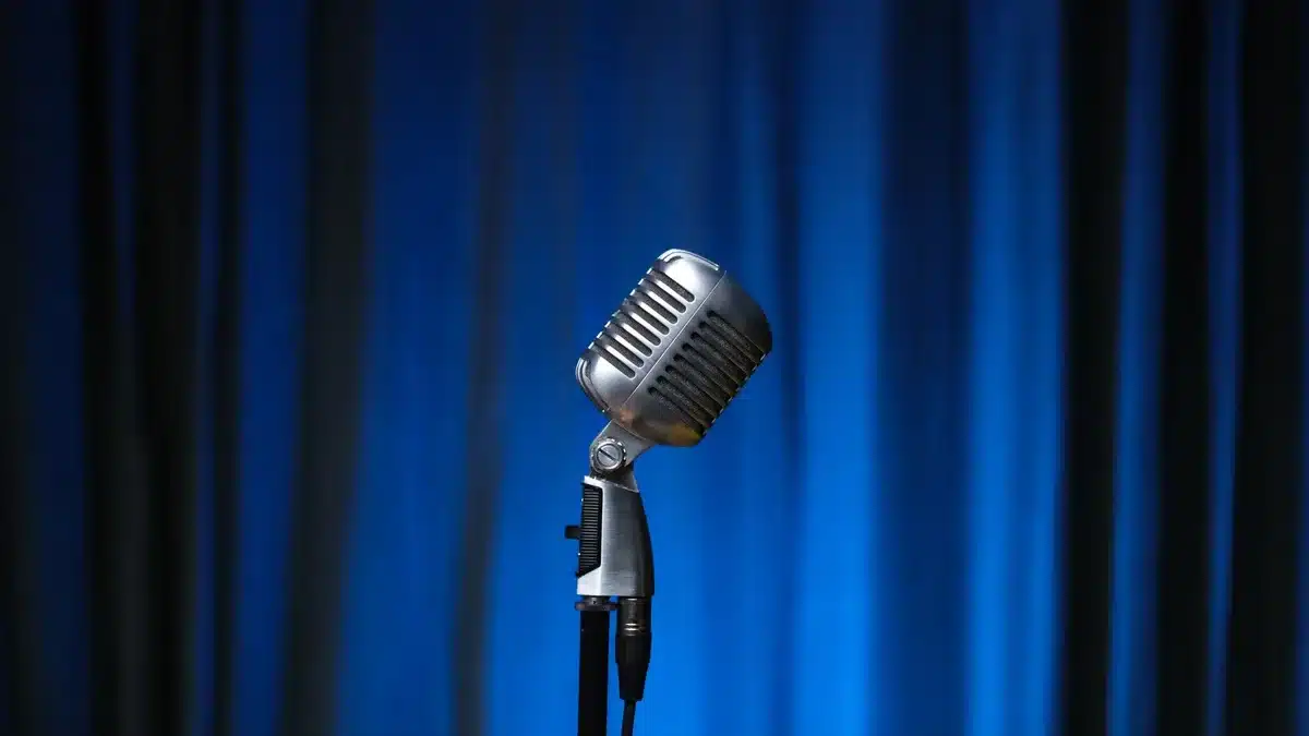 An empty rehearsal stage with a single vintage microphone under a spotlight, scattered papers and a closed notebook on a small table nearby, evoking archival material and memory