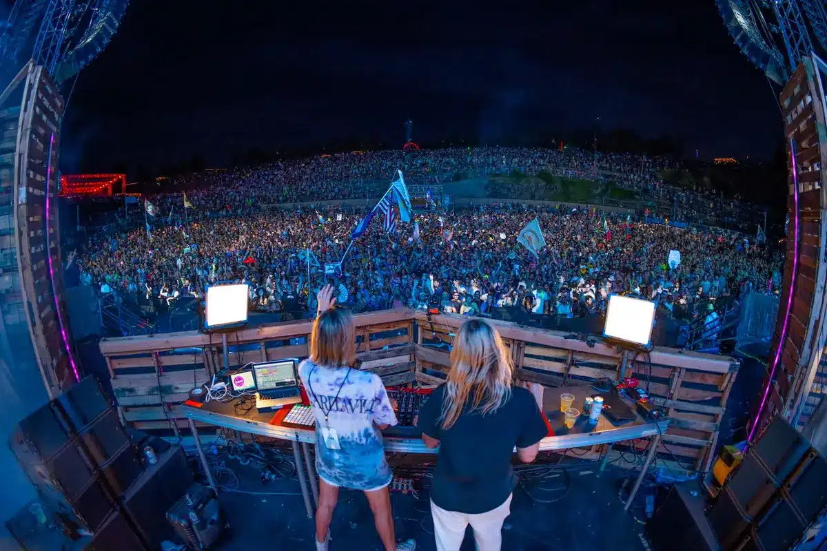 Empty festival field with a large stage structure being assembled, scattered gear and anonymous crew members in the distance; no logos or faces