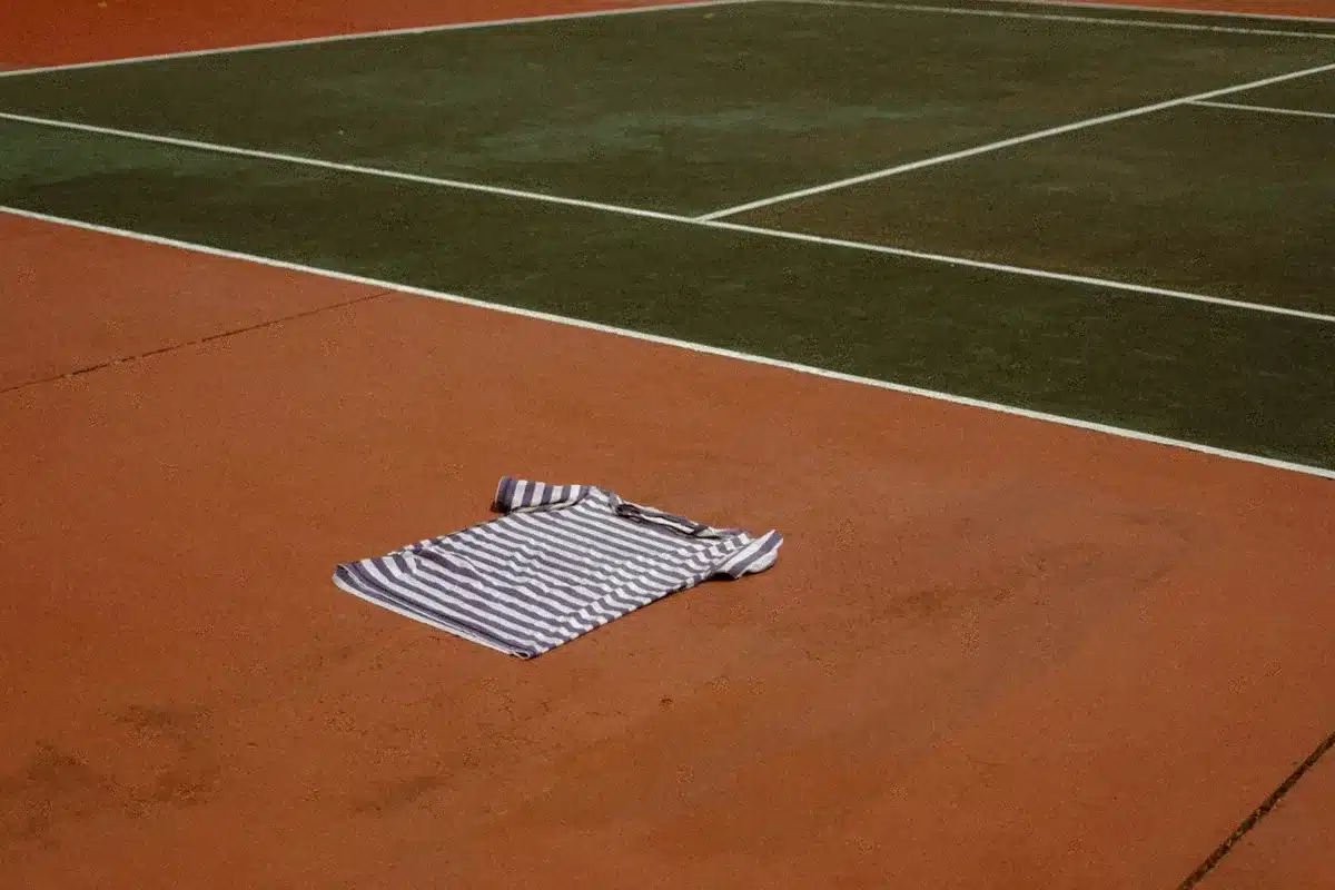 Empty clay court with a lone tennis racket and a single ball near the baseline, dust on the surface, overcast sky above