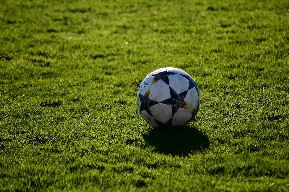 Close-up of a generic match ball on wet turf under stadium lights, empty sideline and a goal net blurred in back