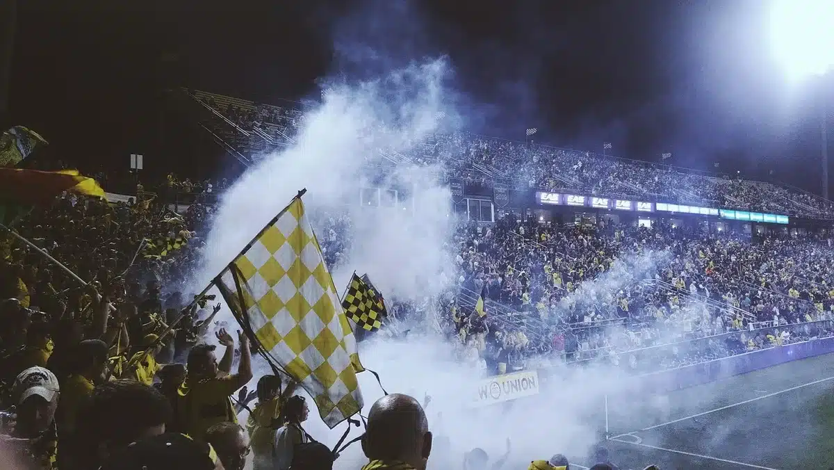 Crowd of anonymous fans outside a generic sports bar watching a big-screen football match; flags and scarves blurred, no logos visible